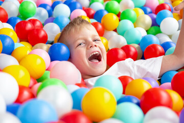 Blonde little boy lying on multi coloured plastic balls in big dry paddling pool in playing centre. Smiling at camera. Portrait close up. Having fun in playroom. Leisure Activity.