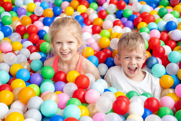 Fototapeta na wymiar Portrait close up Smiling blonde little boy and girl lying on multi colored plastic balls in big dry paddling pool in playing centre. Having fun in playroom Leisure Activity.
