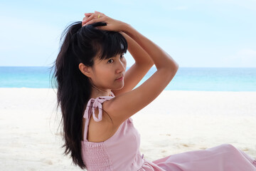 Authentic skin tan asian woman with pink dress sitting on sand beach at coast with blue sea and blue sky blured. she is tourist. summer travel holidays concept.