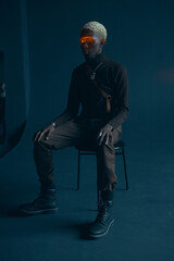 dark-skinned handsome guy with white hair and blue eyes in brown clothes sitting on a chair in a dark studio with a serious expression, where on the eyes shine an orange ray