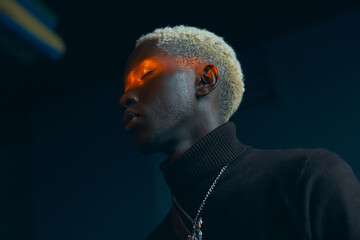 portrait of a dark-skinned handsome guy with white hair dressed in a brown sweater over which hangs a silver chain, he closed his eyes where the orange ray shines