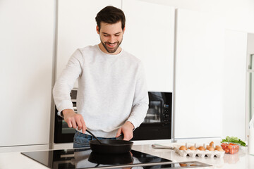 Happy handsome young man smiling while cooking scrambled eggs