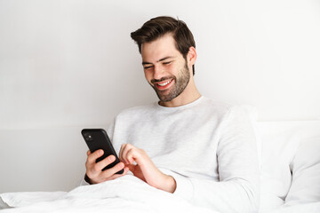 Smiling young man using mobile phone while resting in bed