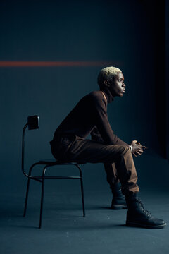 dark-skinned handsome guy with white hair and blue eyes sitting on a chair in a dark studio with a serious facial expression in brown clothes and silver accessories, he looks at the window