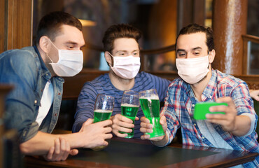 friendship, health and st patrick's day concept - male friends wearing face protective medical mask for protection from virus disease drinking green beer and taking selfie with smartphone at bar