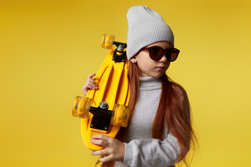 Stylish little child girl in hat and sunglasses posing with yellow skateboard on yellow background