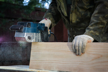 Hands of a joiner with a grinder machine. Flying shavings, wooden board. The work of a professional. Woodwork.