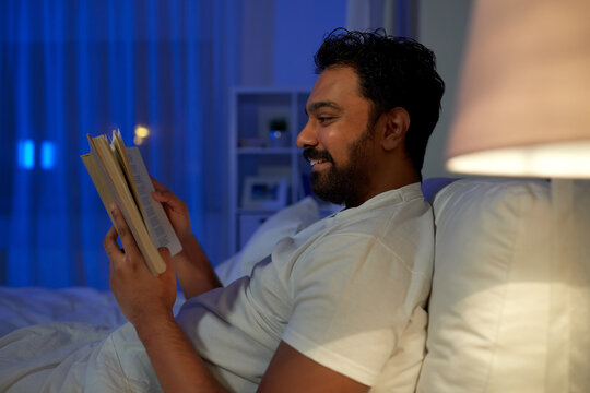 people, bedtime and rest concept - happy smiling indian man reading book in bed at home at night