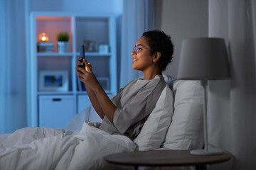 technology, internet, communication and people concept - happy smiling young african american woman in glasses with smartphone lying in bed at home at night
