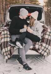 
Couple sitting on a blanket in the trunk of a car and drinking coffee in cups against the background of a pine forest