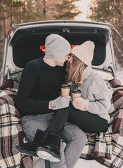 
Couple sitting on a blanket in the trunk of a car and drinking coffee in cups against the background of a pine forest
