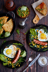Vertical composition. Breakfast of scrambled eggs in the shape of a heart and sausages with vegetables on dark plates on a dark wooden background. View from above.
