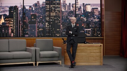Late-night talk show host is leaning against his table in studio, looking into camera. TV broadcast style show