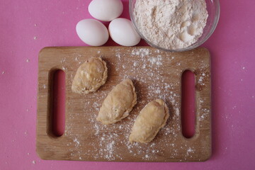 Raw cooking dumplings lie on a board beside egg flour on a pink background view from above
