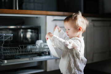 child toddler unloads the dishwasher on his own in a Scandinavian-style kitchen, child's mother's...