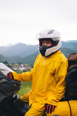 A girl in a yellow raincoat, shoe covers and a motorbike helmet. Motorcyclism and travel. Vertical photo. Top of the Mountains. Close-up view. Difficult test and extreme vacation. Biker outfit.