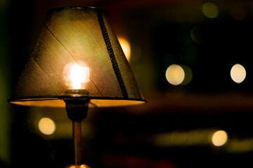 Closeup of a lamp with dark lampshade and warm light in a moody dark room. One lamp on the front...