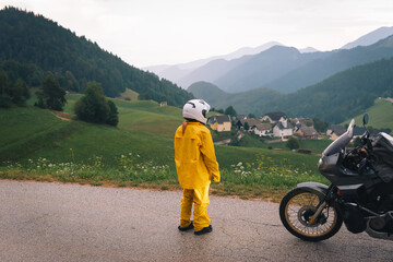 A girl in a yellow raincoat, shoe covers and a motorbike helmet. Motorcyclism and travel. Sightseeing tour. Top of the Mountains. A day with clouds. Difficult test and extreme vacation. Biker outfit.