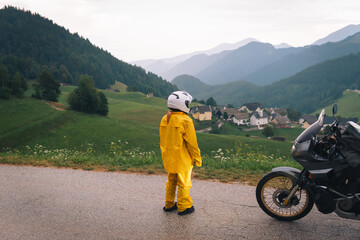 Fototapeta premium A girl in a yellow raincoat, shoe covers and a motorbike helmet. Motorcyclism and travel. Sightseeing tour. Top of the Mountains. A day with clouds. Difficult test and extreme vacation. Biker outfit.