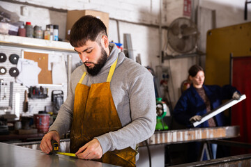 Young bearded man working in industrial workshop, measuring metal structures ..