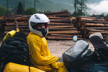 Fototapeta na wymiar A girl in a raincoat, shoe covers and a helmet is sitting on big adventure motorbike. Vacation and travel. Rain and felling after the storm. Sawmill. Difficult road and biker outfit. Close-up