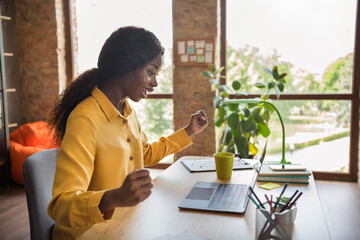 Photo portrait of african american excited girl celebrating working on laptop in modern office indoors