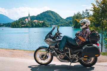 Fototapeta na wymiar Biker man in jeans and a helmet take a rest. Lying on motorcycle with bags. Tourism and vacation. Sunny summer day. Bled lake, island, castle and mountains in background, Slovenia, Europe