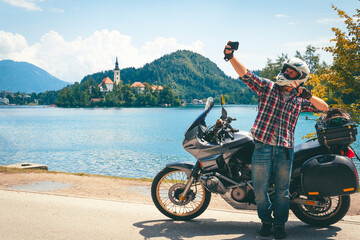 Obraz premium Motorcyclist man takes a selfie on a smartphone. Stands by a motorcycle with bags. Tourism and vacation. Sunny summer day. Bled lake, island, castle and mountains in background, Slovenia, Europe