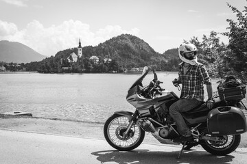 Fototapeta na wymiar Black and white. Rider man in jeans and a helmet. Sits on a motorcycle with bags. Tourism and vacation. Sunny summer day. Bled lake, island, castle and mountains in background, Slovenia, Europe