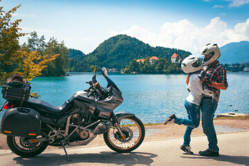 Happy biker couple in love travel together. Stands by a motorcycle with bags. Tourism and vacation. Sunny summer day. Bled lake, island, castle and mountains in background, Slovenia, Europe