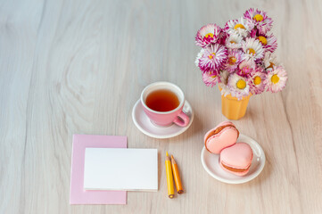  A cup of tea with heart shaped macarons, bouquet of daisies and card for congratulation text on a table. Holiday background, copy space, soft focus, top view