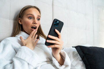 Shocked young woman in bathrobe looking at mobile phone in bed at home