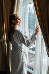Young woman in bathrobe opening bedroom curtains at hotel room