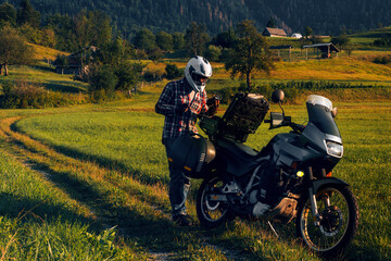 Man motorcyclist Looking for something in the trunk. Alpine mountains on background. Biker lifestyle, world traveler. Summer sunny sunset day. Green hills. hermetic packaging bags. Slovenia