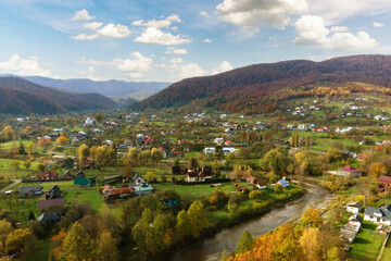Fototapeta premium Aerial view of a rural village with small houses between autumn mountain hills covered with yellow and green pine trees.