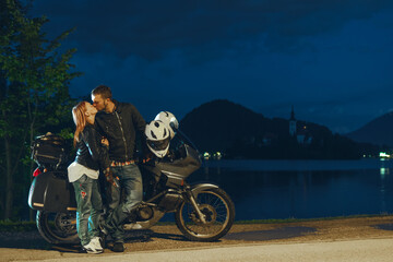 Happy romantic biker couple in love kissing.Motorcycle with bags. Tourism and vacation. Summer night. Bled lake, island, castle and mountains in background, Slovenia, Europe
