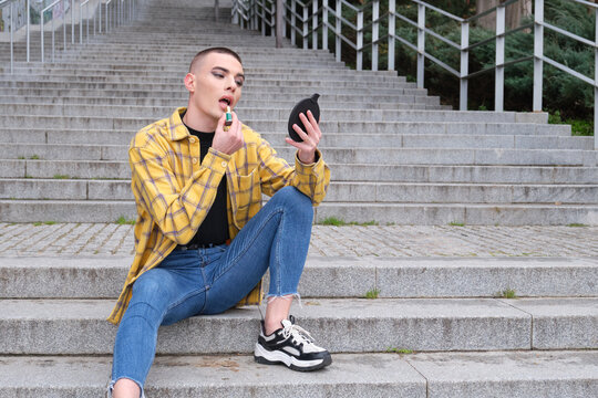 Portrait of a handsome young man, applying lipstick in front of the mirror, wearing black eyeshadow, holding a makeup brush. Non binary androgynous guy.