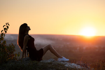 Young elegant woman in black short dress sitting on a rock relaxing outdoors at summer evening. Fashionable female enjoying warm sunset in nature.