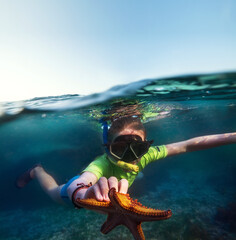 Underwater shot of a young teenage boy snorkeling with dive face mask in the blue Indian Ocean waves near the Zanzibar island with a big orange starfish in his hand. Exotic vacations concept image. - obrazy, fototapety, plakaty