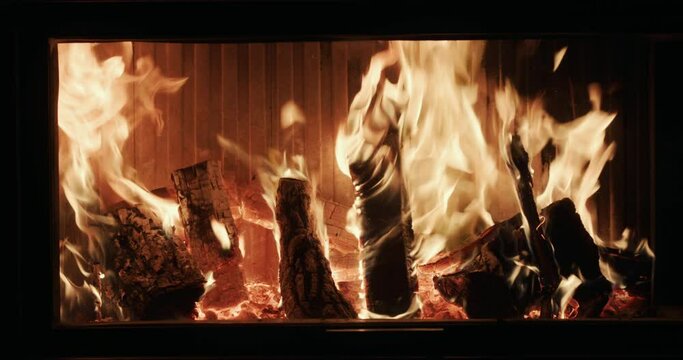 Firewood is burning behind glass in a modern fireplace insert. Slow motion video