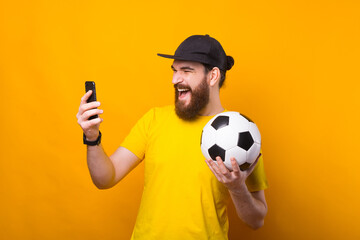 Portrait of amazed bearded young man looking at smartphone and holding soccer ball, support...