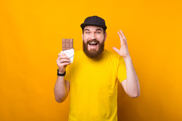Portrait of cheerful young bearded hipster man holding bar of chocolate over yellow background.
