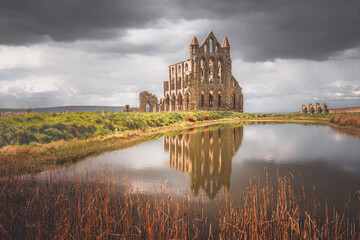 The historic landmark 7th century ruins of Whitby Abbey perched atop East Cliff north Yorkshire, England with a pond reflection against a dark, moody sky.