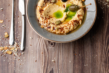 Hummus in a grey bowl seasoned with sunflower and pumpkin seeds, sesame, olives and olive oil. Close-up on a table with cutlery and seeds by side. Top view isolated on wooden background. Copy space.