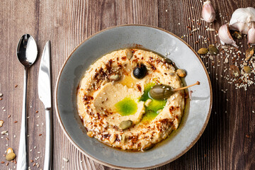 Hummus in a grey bowl seasoned with sunflower and pumpkin seeds, sesame, olives and olive oil. Close-up on a table with cutlery, seeds and garlic by side. Top view isolated on wooden background.