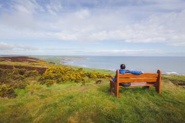 A man enjoys clifftop and seaside landscape views on a park bench at Ravenscar on the North Yorkshire Coast in North York Moors National Park, England.