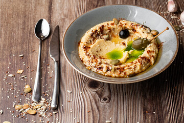 Hummus in a grey bowl seasoned with sunflower and pumpkin seeds, sesame, olives and olive oil. Close-up on a table with cutlery, seeds and garlic by side. Isolated dish on wooden background.