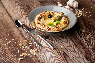 Hummus in a grey bowl seasoned with sunflower and pumpkin seeds, sesame, olives and olive oil. Close-up on a table with cutlery, seeds and garlic by side. Isolated dish on wooden background.