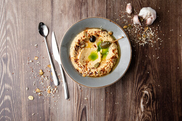Hummus in a grey bowl seasoned with sunflower and pumpkin seeds, sesame, olives and olive oil. Close-up on a table with cutlery and seeds by side. Top view isolated on wooden background. Copy space.