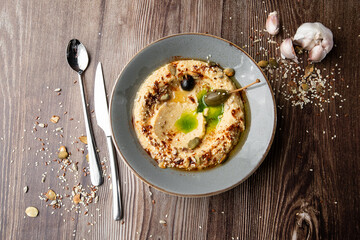 Hummus in a grey bowl seasoned with sunflower and pumpkin seeds, sesame, olives and olive oil. Close-up on a table with cutlery and seeds by side. Top view isolated on wooden background. Copy space.